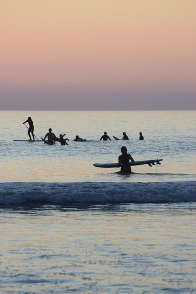Surfer heads into the ocean with demure late fall sunset colors. A paddle boarder is among the surfer line-up