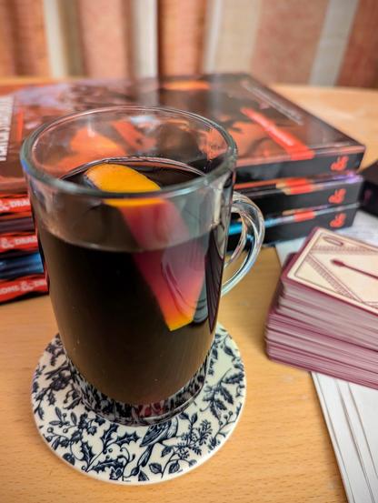 A photo of a red-wine coloured drink in a small glass mug with an orange wedge garnish, sitting on a table alongside various DnD things - three handbooks, some Bard spell cards, and some note paper.