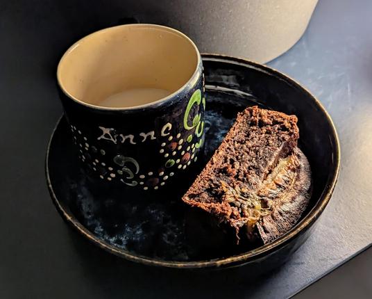 Ein Teller mir hohem Rand in Dunkelblau. Darauf steht eine kleine Tasse mit Blumenmuster, auf der der Name "Anne" steht. Daneben liegt auf dem Teller ein Stück Bananenbrot. Die gebackene Banane auf dem oberen Teil sieht dem Alien aus der Filmreihe "Alien" sehr ähnlich.