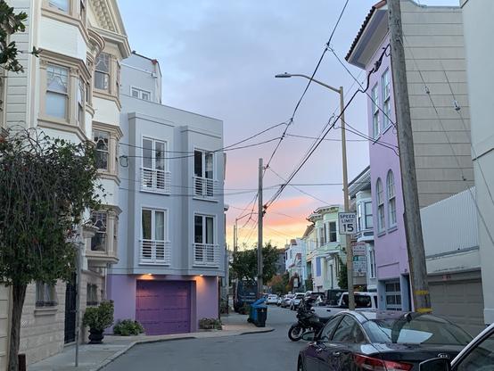 a sunset with yellow sky fading into orange, purple, and gray-blue clouds at the end of a narrow residential street that jogs right. At the jog, two houses flank the street painted in pastel purple on the right, purple ground floor and white upper floors on the left