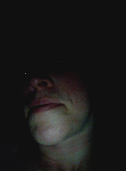A face in the darkness. The neck, chin, lips, and nose are illuminated. The pupils are dots of lights from the flash.