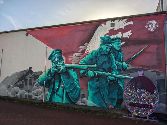 Mural przedstawiający trzech powstańców z karabinami na tle czerwonej flagi z białym orłem. Obok stoi kosz do zbierania nakrętek.