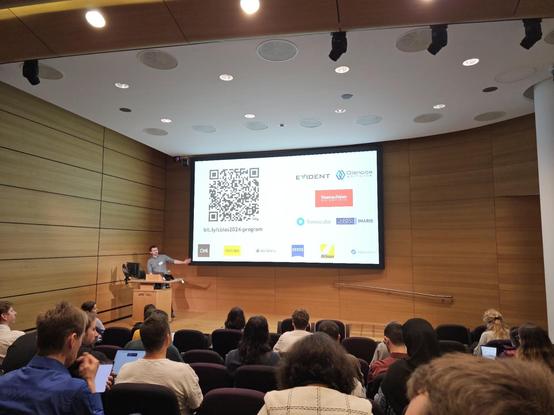 Intro slide for CBIAS 2024 at the Crick. David Barry  is standing in front of the audience and talking about a slide with a QR for the programme.