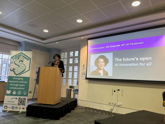 A black woman (Onwurah) stands at a podium with a banner for "The Turing Way" beside them. A slide is projected, featuring the keynote speaker's name and the theme "The future's open - AI Innovation for all."