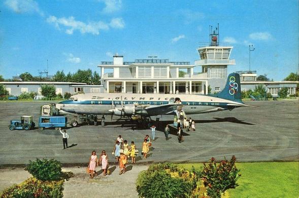Under blue skies, people disembark from a prop plane on the tarmac. Interestingly, they are walking towards the viewer, yet the terminal is in the background.