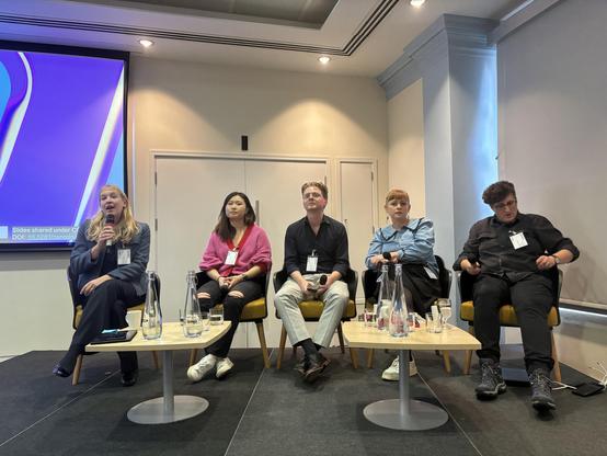 A panel discussion featuring five speakers seated on stage. The host - Aida - is speaking into a microphone, while the others listen. The setting includes a presentation screen in the background and a table with water pitchers and glasses in front of the panelists. There are 4 women and 1 man.