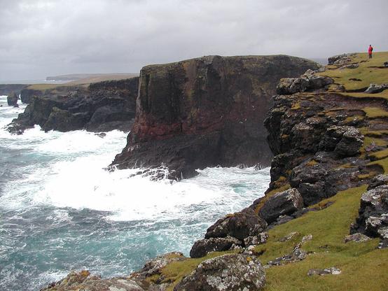 Spectacular cliff scenery near Eshaness lighthouse on the west coast of Shetland on a stormy day. The image shows white waves crashing against dark cliffs, with a single small figure in red standing on top of the cliffs in the upper right of the frame. The cliffs face left and the surface of the sea is largely covered in white foam. The sky is grey.