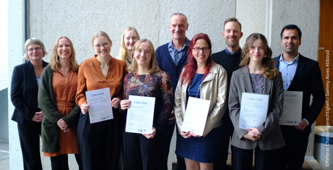 A group of eight people is standing side by side, smiling at the camera. Four people in the front row are holding certificates in their hands. The individuals are dressed in formal attire, and the background is a plain gray wall.