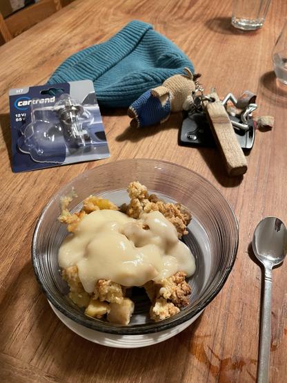 A bowl of dessert with crumbled cake topped with creamy sauce is placed on a wooden table. Nearby, there is a blue knitted hat, a packaged car part, a small stuffed toy, a set of keys, and a glass of water. A spoon is lying next to the bowl.