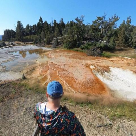 Photo of a person standing on a boardwalk, observing a hot springs area in Yellowstone National Park.