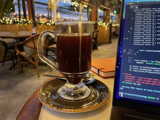 Photo of a bar/restaurant with a sunset visible through the windows at the back. In the foreground is a cup of mulled wine and a partially-visible computer screen showing some failed PlayWright tests.