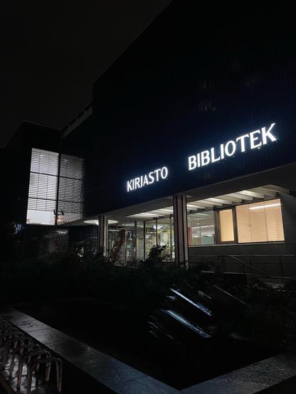 Pimeässä erottuvat kirjaston ääriviivat ja valiokyltit Kirjasto Bibliotek. Etualalla kaide ja istutuksia.