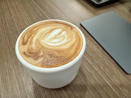 Small white coffee mug filled with a cappuccino sitting on a dark wooden table with a black trackpad sitting near.