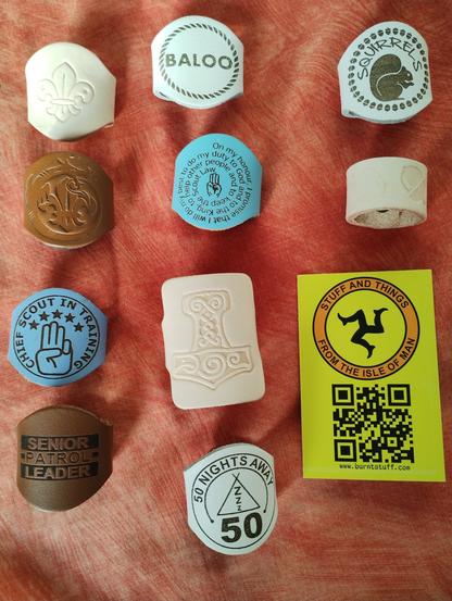 10 leather Scouting neckerchief woggles, 8 of which are circular, 1 a ring loop and the other rectangular.

One has the Christian version of the Scout Promise written in a spiral, one has an embossed Fleur De Lis with a plain design, another a Fleur De Lis with a dragon eating its tale, one says Baloo in a rope ring, another has Squirrels with a squirrel below it, one is a ring of embossed acorns, one is a hand doing the Scout Sign with Chief Scout In Training written around it, another has 50 Nights Away on it. A brown one has Senior Patrol Leader on it and the rectangular one has an embossed Viking rune that looks hammer shaped.

A business card is laid out alongside them.