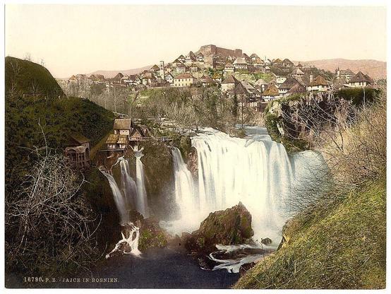 The image depicts a picturesque view with Jajce, Bosnia during the Austro-Hungarian Empire era. It shows two magnificent waterfalls cascading into a river below and is surrounded by lush greenery on hillsides. Traditional houses are visible in the background perched atop the hillside overlooking the falls, creating an idyllic setting that reflects both natural beauty and cultural heritage of the region during this time period.
The date "18790" and location information indicate it was taken around 1890 - ca. 1900 in Jajce, Bosnia under Austro-Hungarian rule. The photo is part of a collection that captures various aspects of life within the empire during this era.