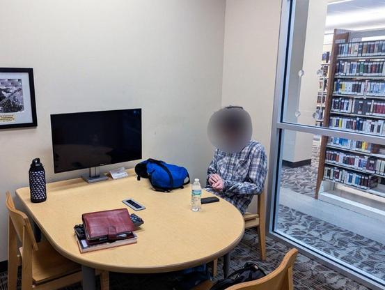 View of a room with a glass wall through which you can see stacks of books 

Inside the room you can see a table with a monitor and a stack of books 

In a chair sits my fiance, with his face blurred for privacy
