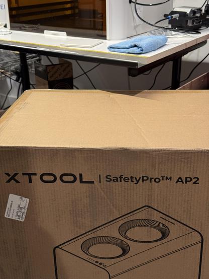 In the foreground: the outer box for an xTool SafetyPro™️ AP2 air purifier. In the background: the riser base for an xTool M1 laser engraver.