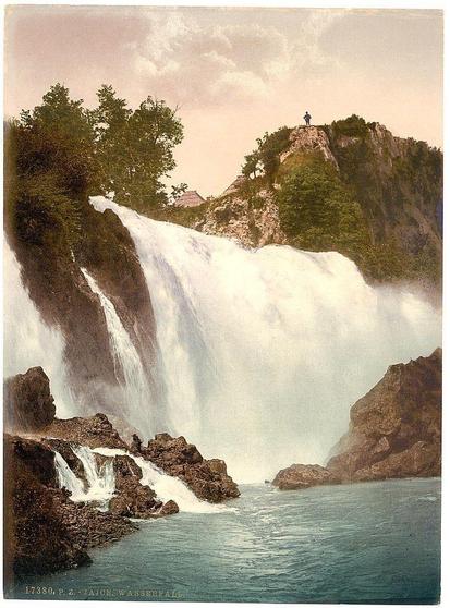 This image depicts a stunning waterfall with multiple cascades flowing down rocky cliffs into the water below. The falls are surrounded by lush greenery and trees, creating a picturesque natural setting. In the background, there is a person standing on top of one of the rock formations, looking out over the scene from above. This photograph appears to be an old colorized postcard or print with some text visible at the bottom left corner indicating "Jajce - Wasserfall" and what seems like a catalog number 17380 P.Z., suggesting it is part of a collection related to Austria-Hungary between approximately 1890-1900. The overall mood conveyed by this image is serene and breathtaking, capturing the beauty of nature's force in its raw form.
