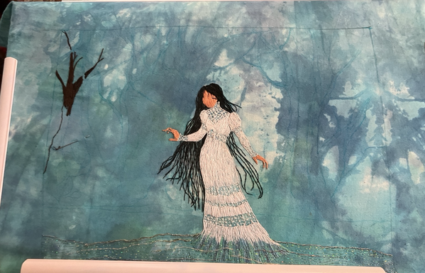 An embroidered panel in progress showing La Llorona standing in the river, with trees sketched into the background and one being filled with dark brown thread.