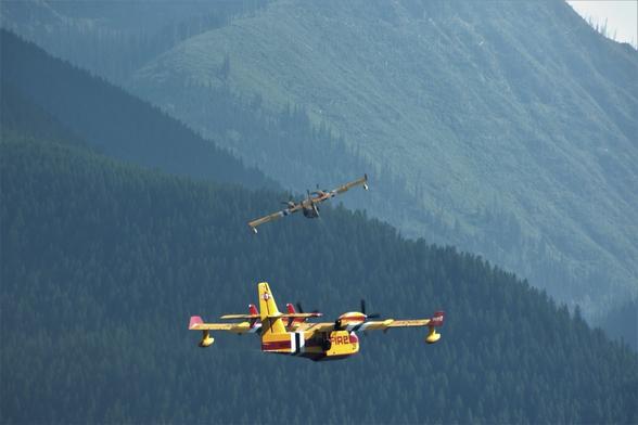 Two yellow airplanes with propellers on them are coming in towards a lake to scoop water for firefighting in the background is a mountain covered with green trees