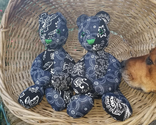 Two cotton teddy bears sitting in a wicker laundry basked which is tipped onto one side to create a photo space booth thing. The bears are made in patchwork out of two fabrics. One fabric is black with a big white floral print and the other fabric is navy blue with a pattern of dots and circles. The bears have green eyes and green noses. On the right hand side of the photo, a  brown dog nose has entered the frame to sniff a bear.