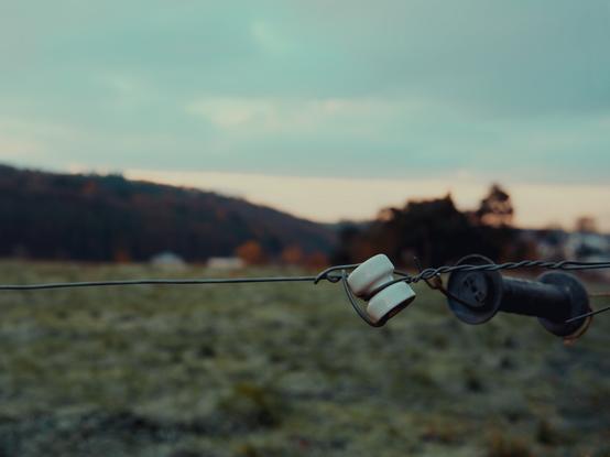 A close-up of an electric fence insulator with two white ceramic rings, set against a soft-focus backdrop of a grassy field and hills in the early morning light. The muted colours of dawn cast a serene, chilly atmosphere, with hints of frost on the ground and a calm sky overhead.