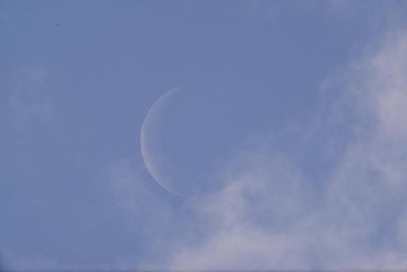 Die abnehmende Mondsichel zwischen Wolkenfetzen am hellen Taghimmel