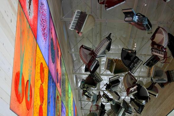 A flock of piano accordions suspended from the ceiling in bellows-unfurled formation swoop past a series of abstract paintings hung near the ceiling of the cafe at the Dale Chihuly glassblowing museum.
