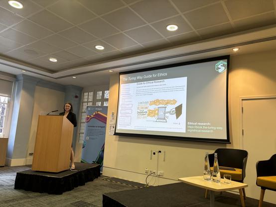 A woman - Arielle - stands at a lectern giving a presentation on ethical research, with a visual guide titled "The Turing Way Guide for Ethics" displayed on a screen. The background features signs promoting UK AI innovation. Two empty chairs and glasses of water are visible