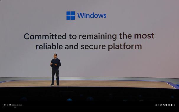 A screenshot of Microsoft's keynote, with a person in front of a giant screen that says "Windows, committed to remaining the most reliable and secure platform"