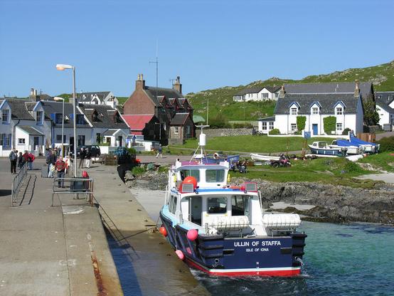 The ferry slipway and the village of Baile Mor on Iona, shown on a sunny day with clear blue skies. In the lower left of the frame is a concrete slipway and a blue and white boat is moored to it, facing away from the camera. It is called Ullin of Staffa. The village is in the background, mainly comprising white buildings, though one is of stone. The land rises in the background.