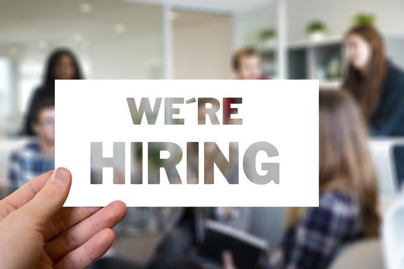 Eine weiße Karte wird von einer Hand hochgehalten. "We're Hiring" steht in großen Buchstaben darauf. Im Hintergrund sieht man unscharf Menschen und Regale