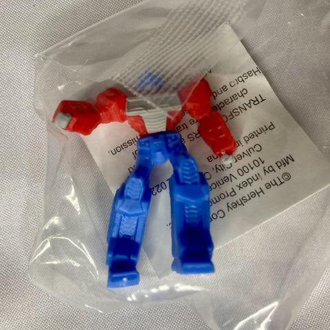 A small, toy action figure with a red upper body and blue lower body is seen partially inside a transparent plastic bag. A white tag with text is visible beneath the figure.