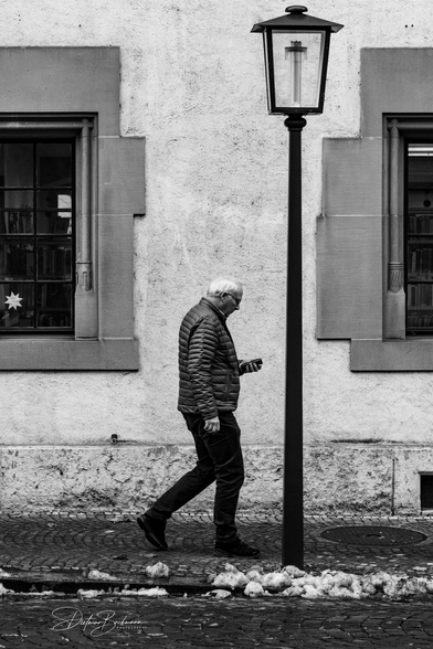 Das Schwarz-Weiss-Foto zeigt einen älteren Mann, der in einer gepolsterten Jacke und Jeans eine gepflasterte Strasse entlangläuft. Er schaut konzentriert auf ein Smartphone in seiner rechten Hand, während er scheinbar direkt auf einen Laternenmast zuläuft. Im Hintergrund ist eine grob verputzte Wand mit zwei Fenstern zu sehen, die von steinernen Rahmen eingefasst sind. Zwischen den Fenstern steht die nostalgisch anmutende Laterne, die dem Mann im Weg zu sein scheint. Im unteren Bereich des Bildes sind Reste von schmutzigem Schnee am Strassenrand sichtbar. Das Bild strahlt eine ruhige, alltägliche Szene aus, eingefangen in klaren Kontrasten.