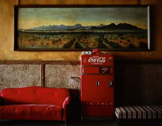 Un salon, avec à droite un canapé rouge (vue partielle) et à gauche une sorte de lit une place. Entre les deux, un distributeur de Coca-Cola. Au mur, un tableau panoramique, un paysage rural étatsunien vide et triste. Tout est vintage, un peu hors du temps.