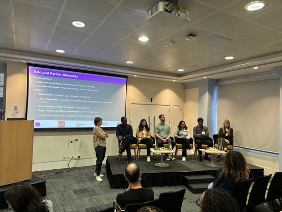 A panel discussion titled "BridgeAI Partner Showcase" featuring six speakers seated on a stage in a conference room. A woman - Dominica - is hosting the session and standing in front of the screen. A screen in the background displays names of panel organizers and speakers, with highlighted roles related to AI and business.