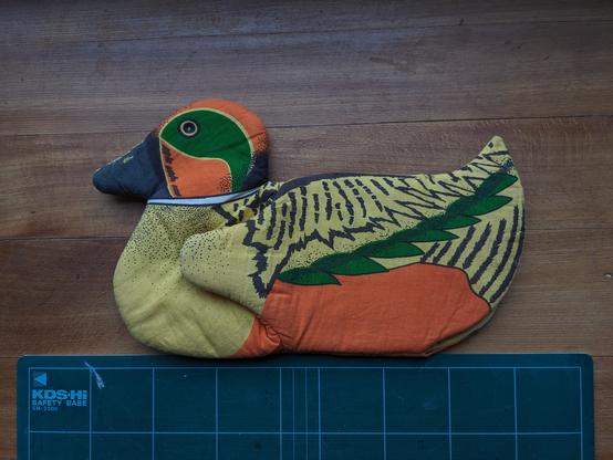 Posé sur une planche, au-dessus d'une planche à découper en plastique canard, une manique canard. Elle est faite d'un tissu imprimé jaune, orange, vert et noir et ressemble à un canard, l'aile dissimule le pouce de la moufle.