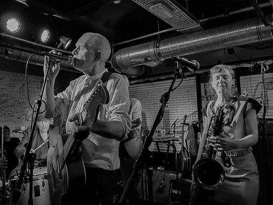 Foto van De Kift live aan het werk, in zwart-wit. Gitarist links, saxofoniste rechts, drummer op de achtergrond.