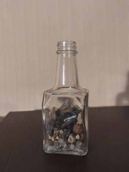 A squareish vinegar bottle without a lid is filled with stones of different sizes, and a single metal screw.