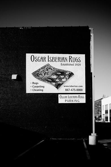 The image shows the exterior of Oscar Isberian Rugs shop. It is a clear day with vivid blue skies, casting bright light on the street and the store.