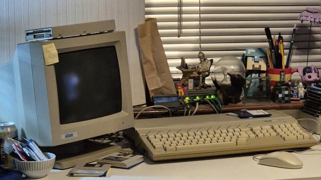 An Atari CRT monitor with external floppy disk drive resting on top sits next to an Atari STe computer on a desk. A network switch and various geeky trinkets sit on the windowsill behind. There are floppy disks, Minidiscs and a stack of hard drives visible to the sides.