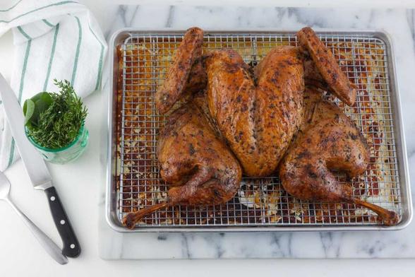 Image of a roasted spatchcocked turkey on a rack over a half-sheet pan. Beside the pan are some herbs in a glass, a carving knife and a white and green tea towel.