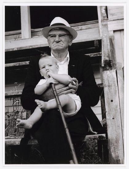 The image is a black and white photograph featuring an older man wearing glasses, a hat, suit jacket, collared shirt, and trousers. He holds a young child with curly hair on his lap while sitting outside against the backdrop of what appears to be part of a porch or wooden structure. The elderly gentleman has one foot resting casually atop the railing behind him, suggesting he is seated comfortably in an outdoor setting.

The older man's facial expression seems contemplative and thoughtful as he looks directly at the camera with a slight frown on his face. His left hand gently grasps a walking cane that rests against the wooden structure beside him. The child appears to be wearing a white top, but their lower body is not fully visible in the frame.

The photograph evokes a sense of nostalgia and captures an intimate moment between the elderly man and the young child, possibly depicting a familial bond or relationship.