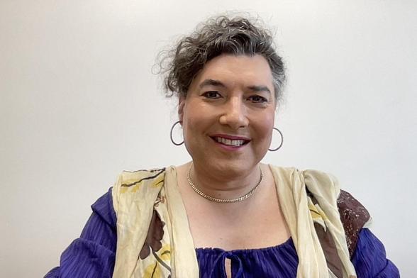 A woman smiling at the camera, wearing a purple dress, a tan, yellow and brown scarf, earrings and a necklace.