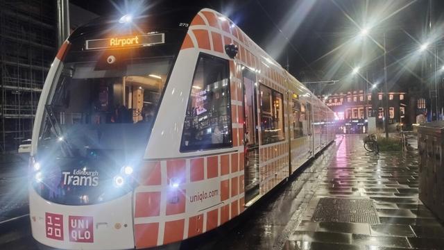 Looking at a slight angle from the front and down the length of an Edinburgh tram, paused at a stop on a wet night, streetlights reflecting off damp pavements