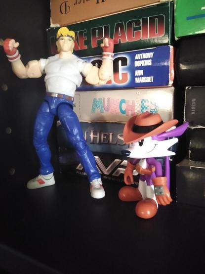 Photo of the Fang action figure standing next to the Axel Stone figure in front of a stack of VHS tapes