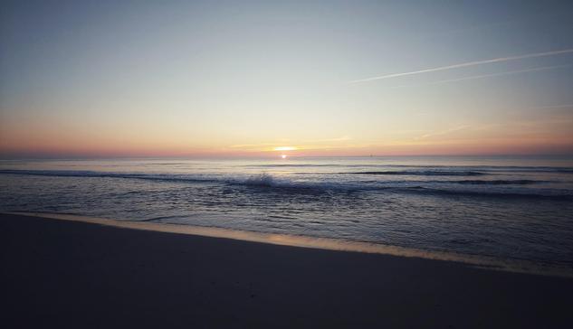 Das Bild zeigt eine Szene am Meer, im Vordergrund ist der Strand zu sehen, er ist menschenleer, die See ist relativ ruhig, lediglich ein paar Brandungswellen wabern in Richtung Strand. Das Wasser glitzert dezent. Am Horizont ist noch ein kleiner Teil der untergehenden Sonne zu sehen, es sieht so aus als würde sie mitten im Meer versinken. Der Himmel ist wolkenlos, lediglich ein paar kleine Wölkchen finden ihren Platz. Die Szene wirkt ruhig, fast still.