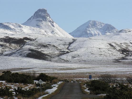 A wintry view of the single track road east from remote Achiltibuie. The image shows a road leading out from the centre of the bottom of the frame with a passing place sign to its left. The road disappears over a hum, leaving snowy land extending out to the start of mountains, which climb to a spiky peak in the top left of the frame and are covered in snow. The scene is in sunlight.
