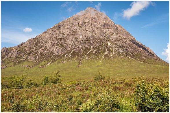 A large, rocky mountain stands prominently against a blue sky with a few clouds. The foreground features lush green vegetation and scattered trees.