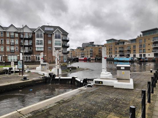 A view of the locks at Brentford Dock, with apartment buildings behind.  The water is swollen to the top of the lock gates, and is spilling through the runnels cut into the tops of the things.
